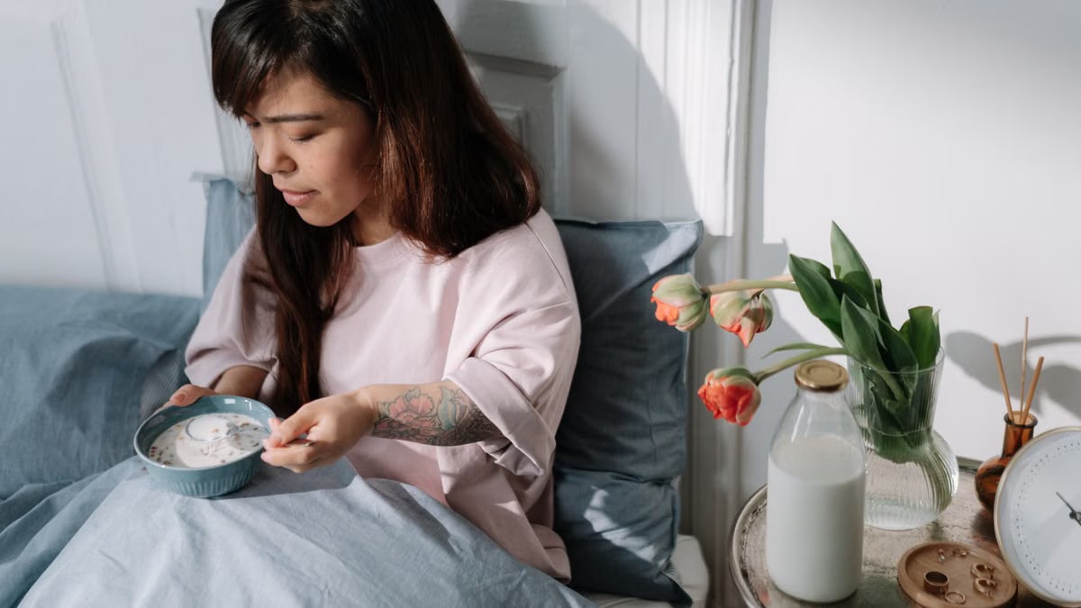 A woman sits in bed enjoying breakfast with milk and flowers nearby, bathed in morning light.